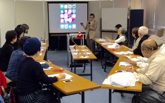 【写真】やまねさんがシニア向けにあいふぉんの講座を開催している。参加者はやまねさんを囲んで真剣に話を聞いている。