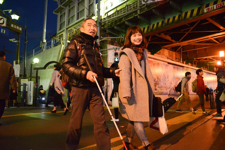 【写真】白杖を持って歩くこばやしこういちろうさんとライターのくどうみずほ