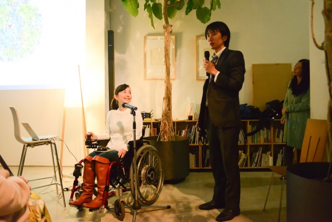 【写真】おださんの隣で参加者にむけて話をするおださんの旦那さんであるよういちさん。おださんは笑顔で旦那さんを見つめている。
