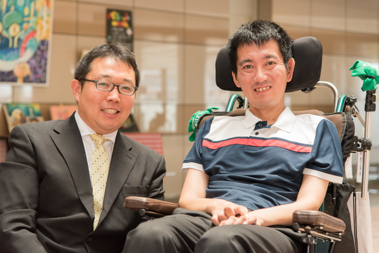 【写真】車椅子に座り笑顔のおんださとしさんとスーツを着ている秘書の坂田さん