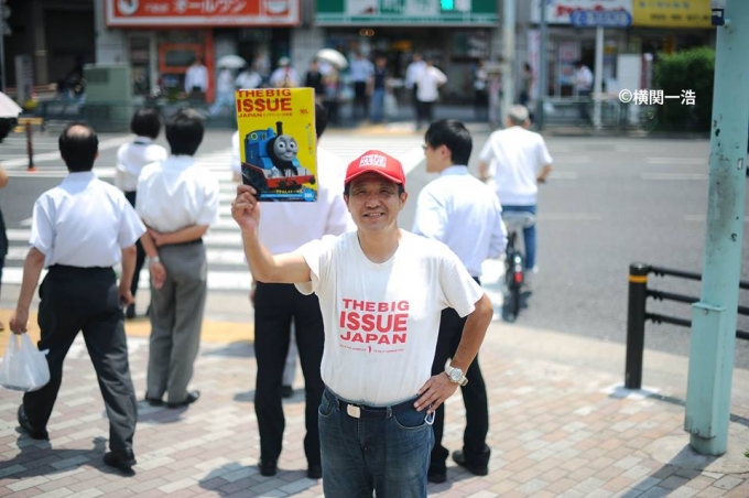 【写真】びっぐいしゅーの雑誌を右手に掲げて笑顔で道にたたずむびっぐいしゅーの販売員