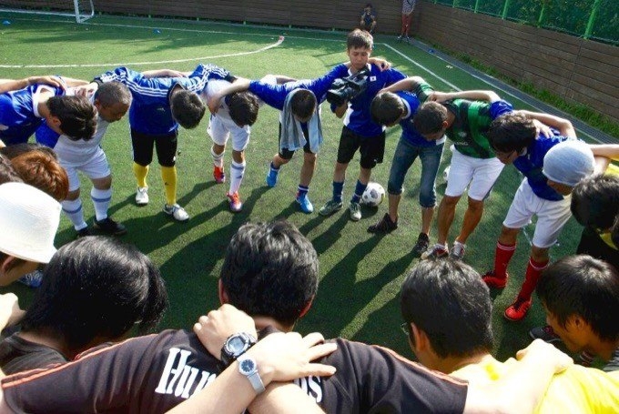 【写真】サッカーのユニフォームを着て大きい円陣を組んでいるチームメンバー