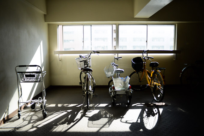 【写真】自転車置き場に並んでいる自転車とお買い物用カート。それらの影が地面に色濃く映っている。