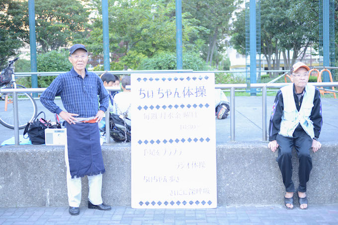 【写真】ちぃちゃん体操の開催時間が書かれた看板の横に座っているスタッフと利用者。体操がはじまるのを一緒に待っている。