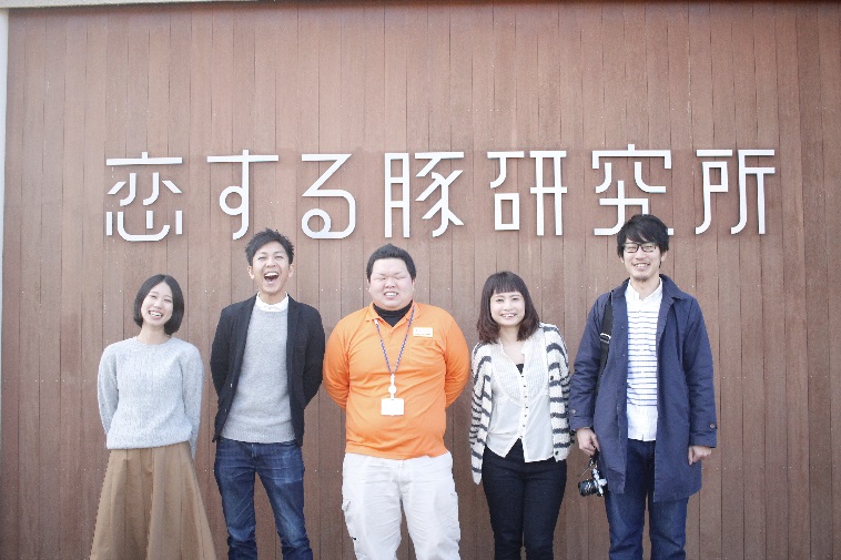 【写真】こいするぶたけんきゅうじょのロゴの前で、取材メンバー3人と、ありたさん、西山さんが並んで笑顔を向けている