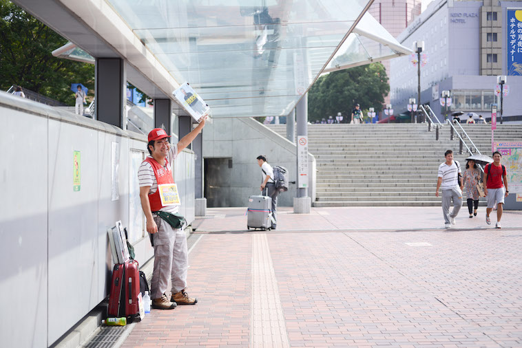 【写真】多摩センター駅前の路上でビッグイシューを掲げるやましたさん