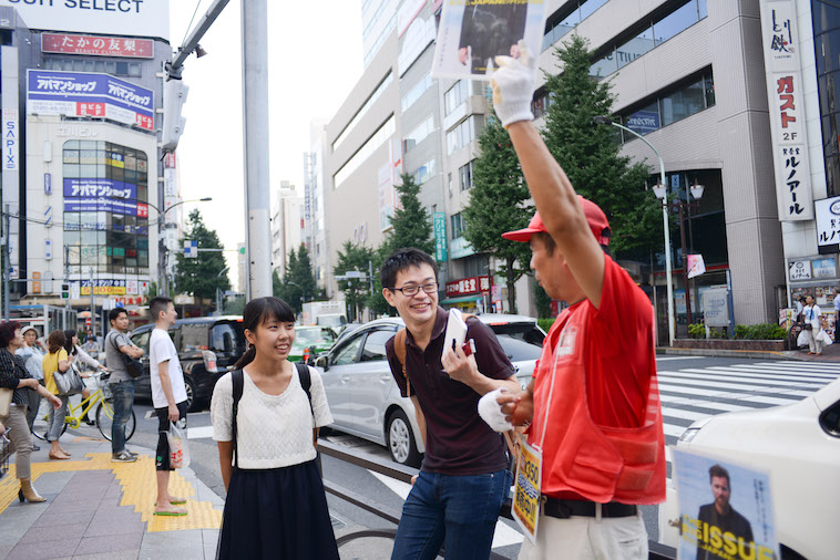 【写真】路上で高々とビッグイシューを掲げている山崎さんとライターのにしやまたけしさん