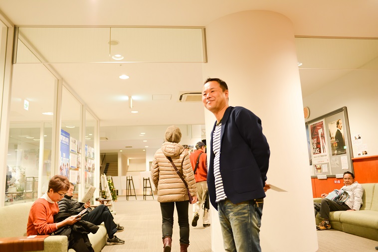 【写真】映画館のロビーでお客さんを待ち佇んでいるあおやまさん。笑みを浮かべている。