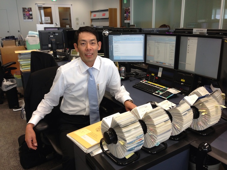 【写真】会社の机に座り、ネクタイをしててこちらに笑顔を向けるおおくぼさん。