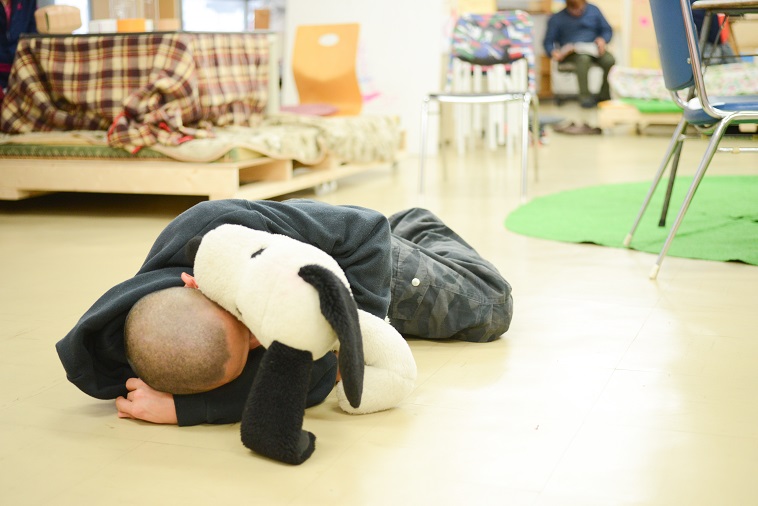 【写真】スヌーピーのぬいぐるみを抱き抱えて、床に寝そべっている利用者。心地よさそうだ。