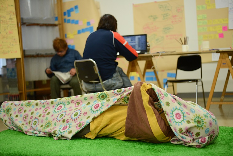 【写真】緑のマットの上に仰向けで寝転がっている利用者。ブランケットを被っている。