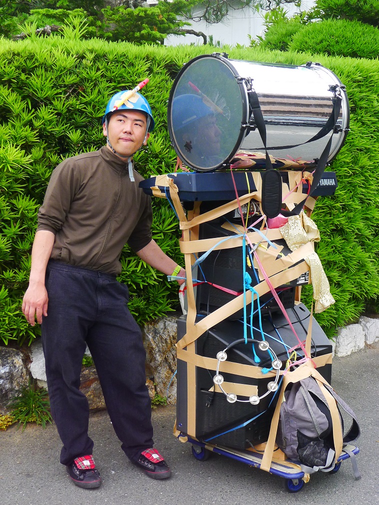 【写真】自身と同じくらいの高さになっている「おが代車」の横でポーズを決めている利用者のおがちゃん。台車にはテレビやドラム、キーボードなどが積み重ねられている。