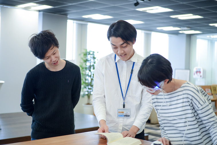 【写真】オトングラスをつけて本を読んでいるとしきよさんと、説明しているしまかげけいすけさん、ライターのこやまかずゆきさん