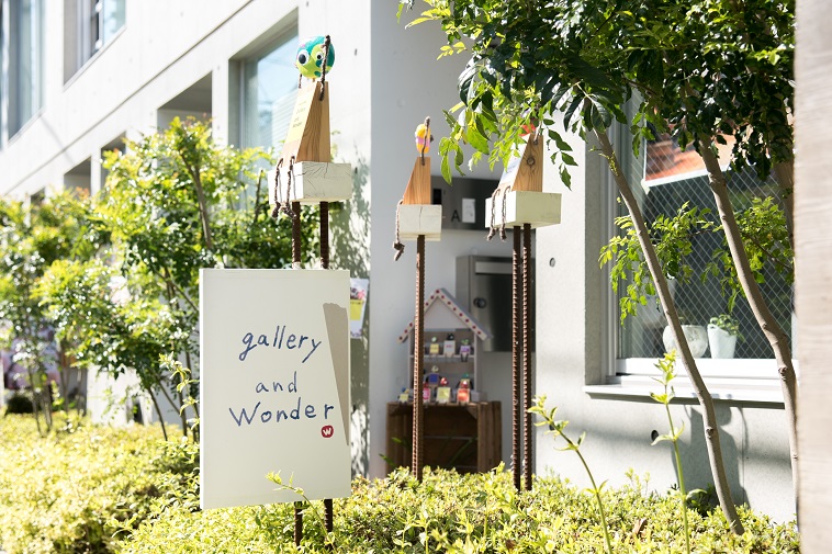 【写真】ギャラリーアンドワンダーと書かれた看板。日光が当たっていたり、緑の木々があり、暖かい空気感がある。