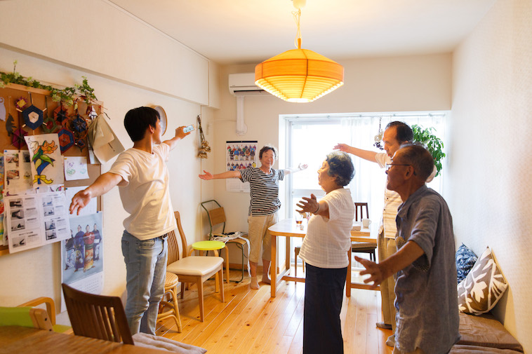 【写真】あたたかな雰囲気の室内で、おじいさんとおばあさんがスタッフとともに体操を行なっている