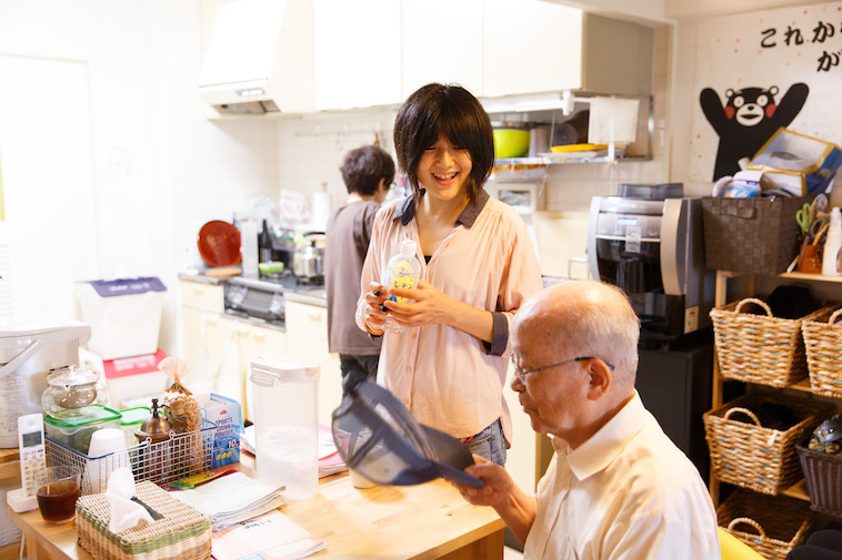 【写真】キッチンスペースに腰掛けるおじいさんに、笑顔で話しかけるたかすさん