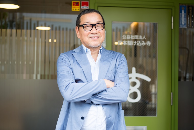 【写真】オフィスの前で腕を組んで笑顔を向けるげんひでもりさん