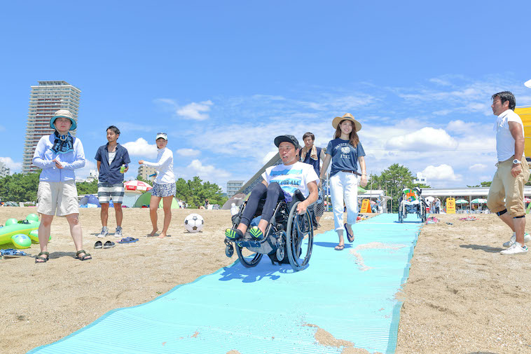 【写真】砂浜でビーチマットの上を歩くきどあやさんと車椅子に乗って移動しているきどしゅんすけさん