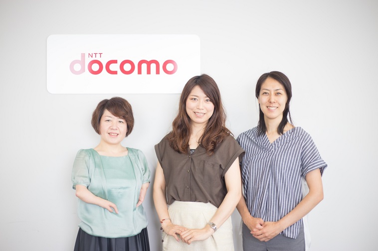 【写真】docomoの看板の前で笑顔で立っている3人の女性