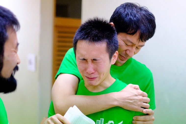 【写真】泣きそうになる高校生の男の子を抱きしめるチームメンバーの男性と、それを微笑みながら見守るもう一人のチームメンバーの男性