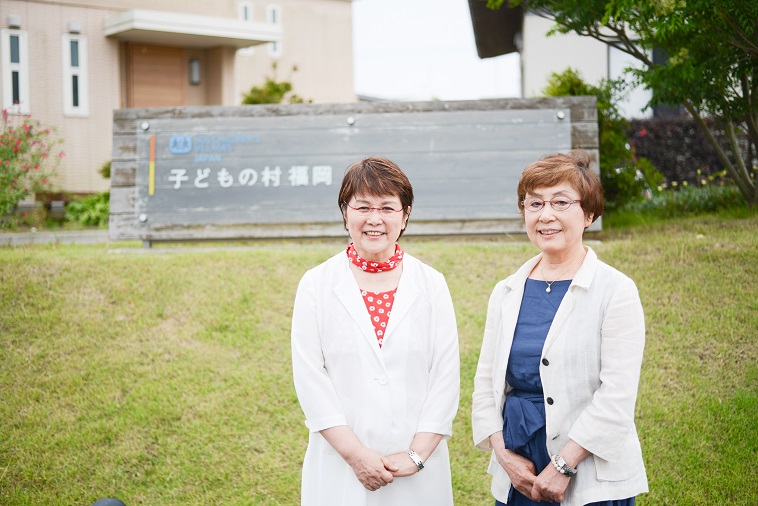 【写真】「子どもの村福岡」の看板の前で笑顔で立っているさかもとまさこさんとおおたにじゅんこさん