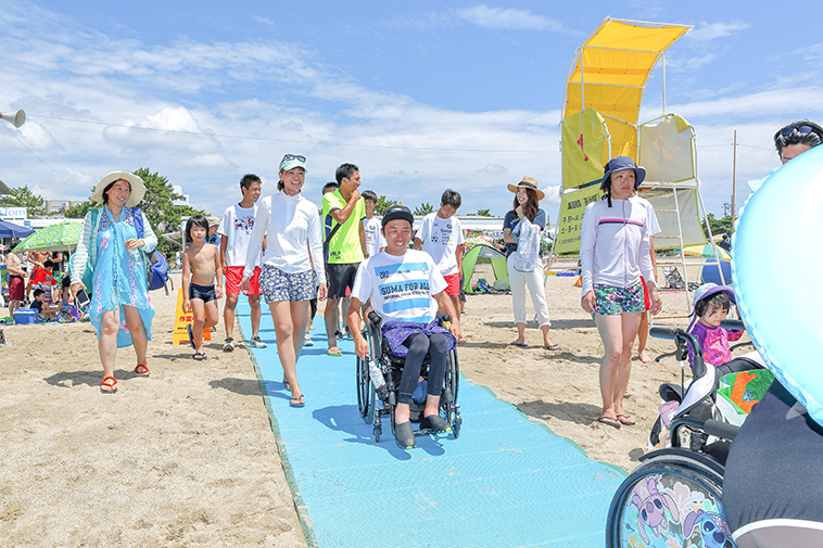 【写真】砂浜に敷いたビーチマットの上を、車椅子に乗った人々が次々に進んでいく。周囲の人々が笑顔で見守っている