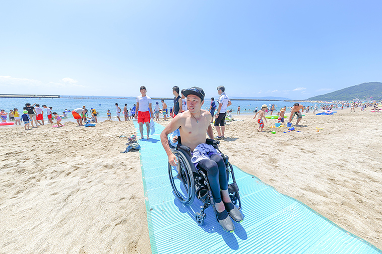 【写真】砂浜に敷いたビーチマットの上を車椅子で進むきどさん