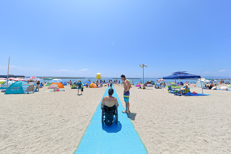 【写真】海に向かってまっすぐ繋がっている砂浜の上のビーチマットを、車椅子に乗った男性が進んでいく