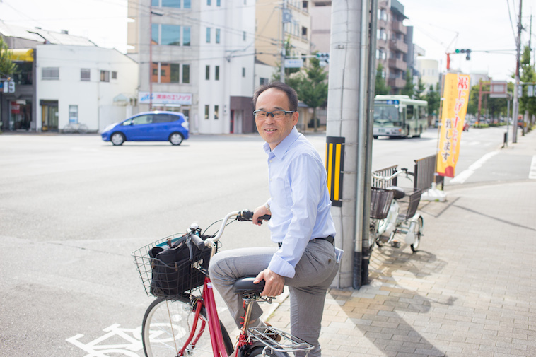 【写真】街頭で笑顔で自転車に乗っているきもとつとむさん