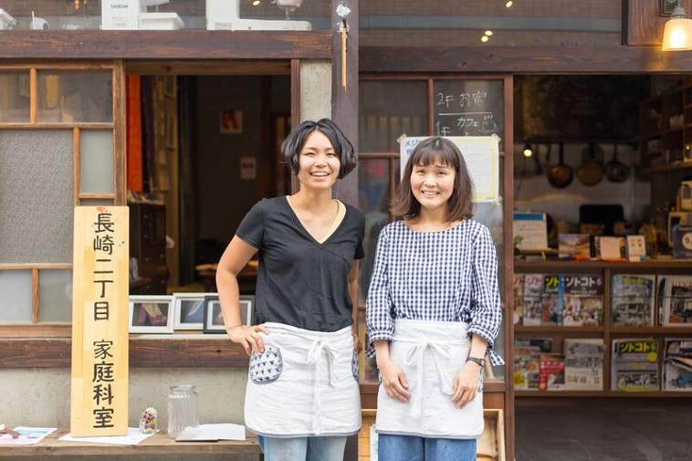 【写真】ながさきにちょうめかていかしつの看板のよこで笑うふじおかさんとふかのさん