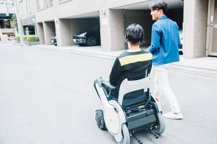 【写真】街頭で車椅子に座って移動するむとうまさたねさんとライターのすずきゆうへい