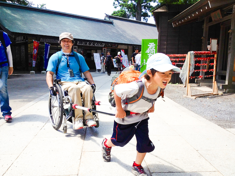 【写真】JINRIKIを使って大人の乗った車椅子を引っ張る少年