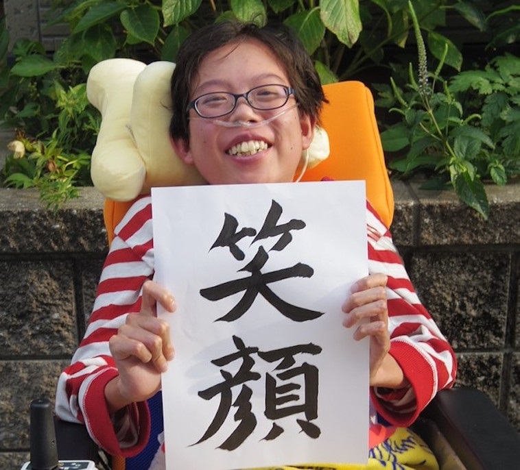 【写真】笑顔という紙を掲げて微笑む車椅子の子ども