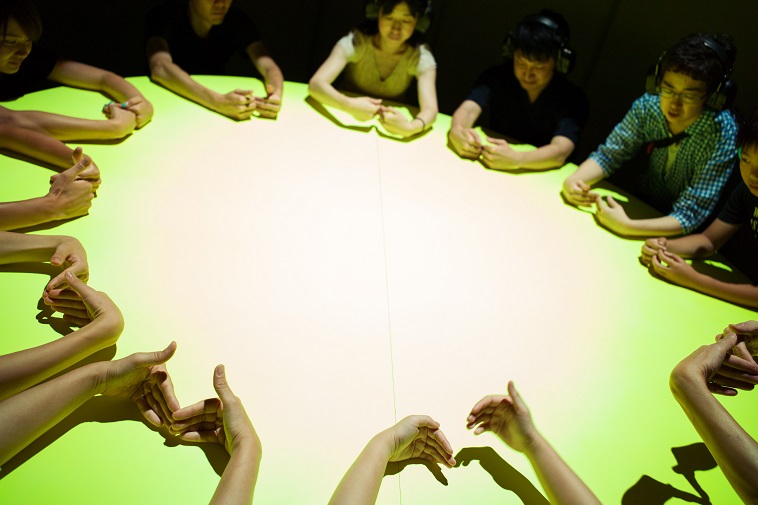 【写真】丸い机を囲み、参加者たちが手遊びをしている。