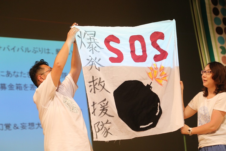 【写真】べてるメンバーが、「爆発救援隊　SOS」の文字と爆弾の絵が描かれた旗を持っている