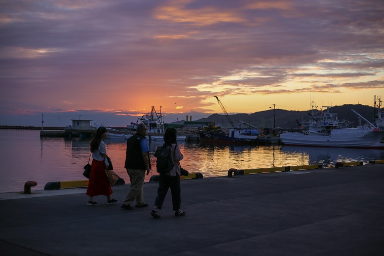 【写真】浦河町の海沿いの道を歩く、はやさかきよしさんとsoar取材チーム。バックには夕日が沈んでいく美しい情景が