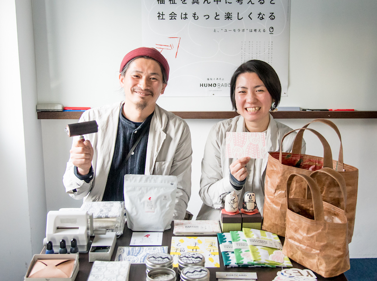 【写真】バッグや手ぬぐい、ポストカードなどデザイン制作した商品を並べた机を前に微笑むHUMORABOまえかわゆういちさんとまえかわあきこさん
