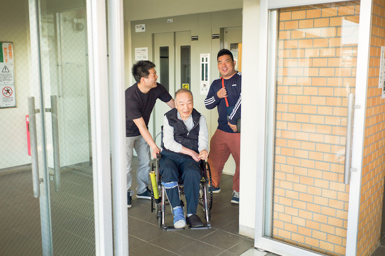 【写真】団地内にあるエレベーターの前で、車椅子に乗る利用者と会話しながら歩くスタッフ。楽しそうだ。