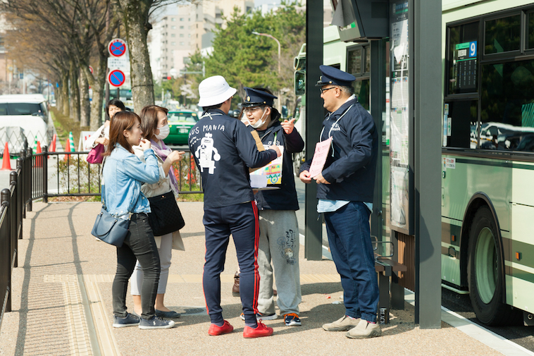 【写真】バス停で交通案内をするメンバーのQさんとXLさん。駅員のような帽子をかぶっている