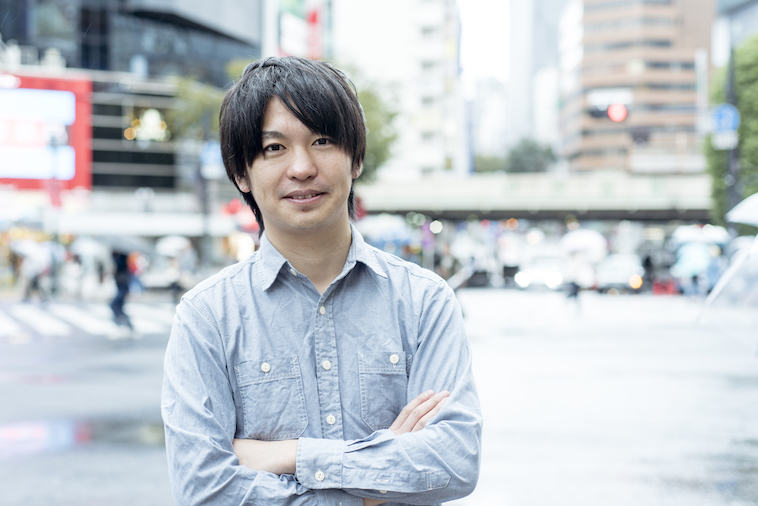 【写真】交差点の前に立ち、爽やかな笑顔でこちらを見るいまいさん。