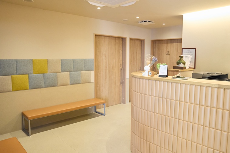 【写真】ゆうりんクリニックの内部。右側に受付があり、左側にベンチが置かれている。オレンジ色の照明で温かな雰囲気がある。