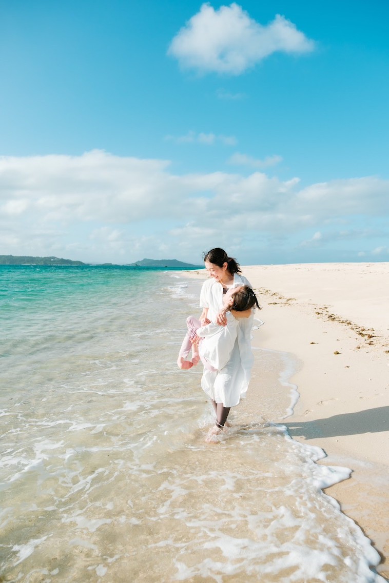 【写真】綺麗な青の海辺の砂浜で、さくらさんがマコさんを抱っこして歩いている