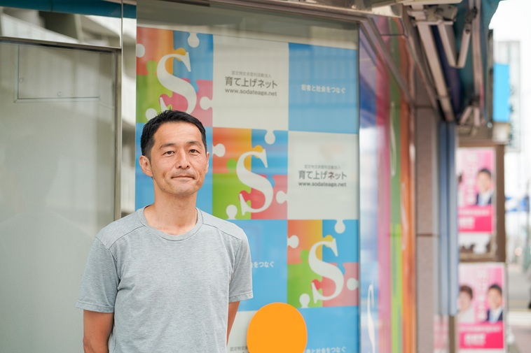 【写真】建物の前でカメラを見つめて微笑むくどうけいさん